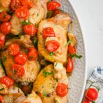 Cooked chicken thighs topped with cherry tomatoes and parsley on white background.