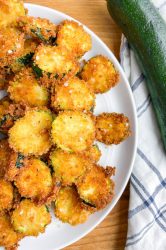 Up close shot of fried zucchini on a white plate with blue and white towel and whole zucchini.