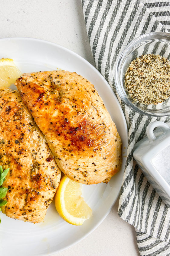 Overhead photo of lemon pepper chicken on a white plate with a lemon wedge, arugula. Striped towel with lemon pepper and salt on the side.