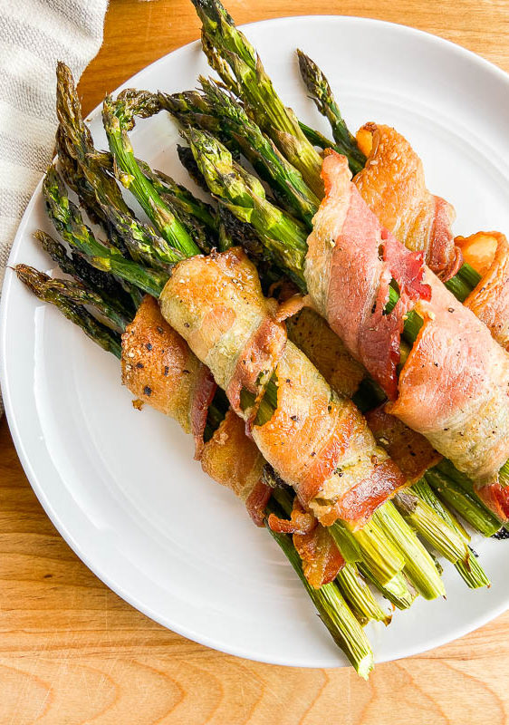 Asparagus wrapped in bacon on a white plate with a stripped towel on a wooden cutting board.