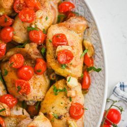 Cooked chicken thighs topped with cherry tomatoes and parsley on white background.