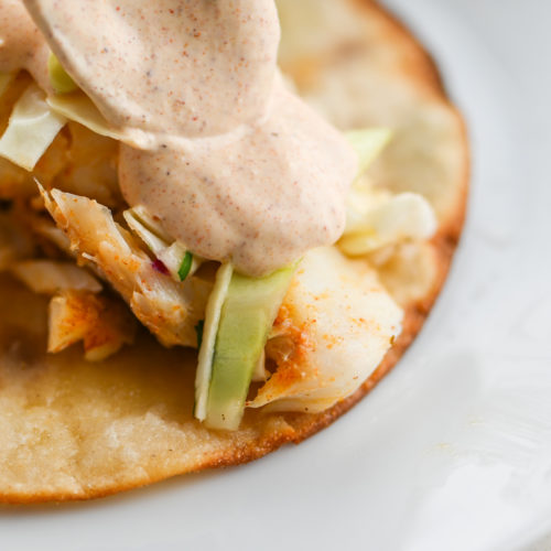 Spooning creamy taco sauce onto fish taco on white plate.