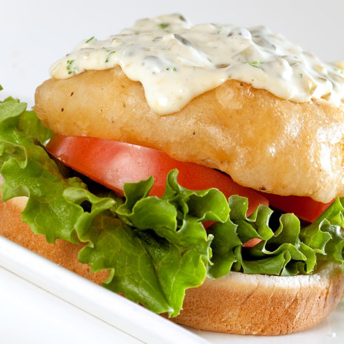 Close up of beer battered fish sandwich on white plate.