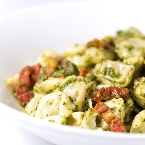 Cheese tortellini with pesto and sundried tomatoes in white bowl.