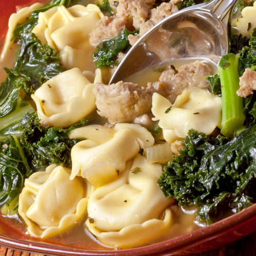 Sausage, kale and tortellini soup in red bowl with a spoon.