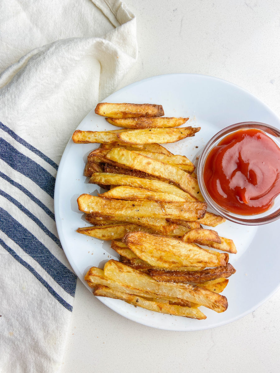 Air fryer french fries on a white plate with ketchup, a striped napkin and white background. 