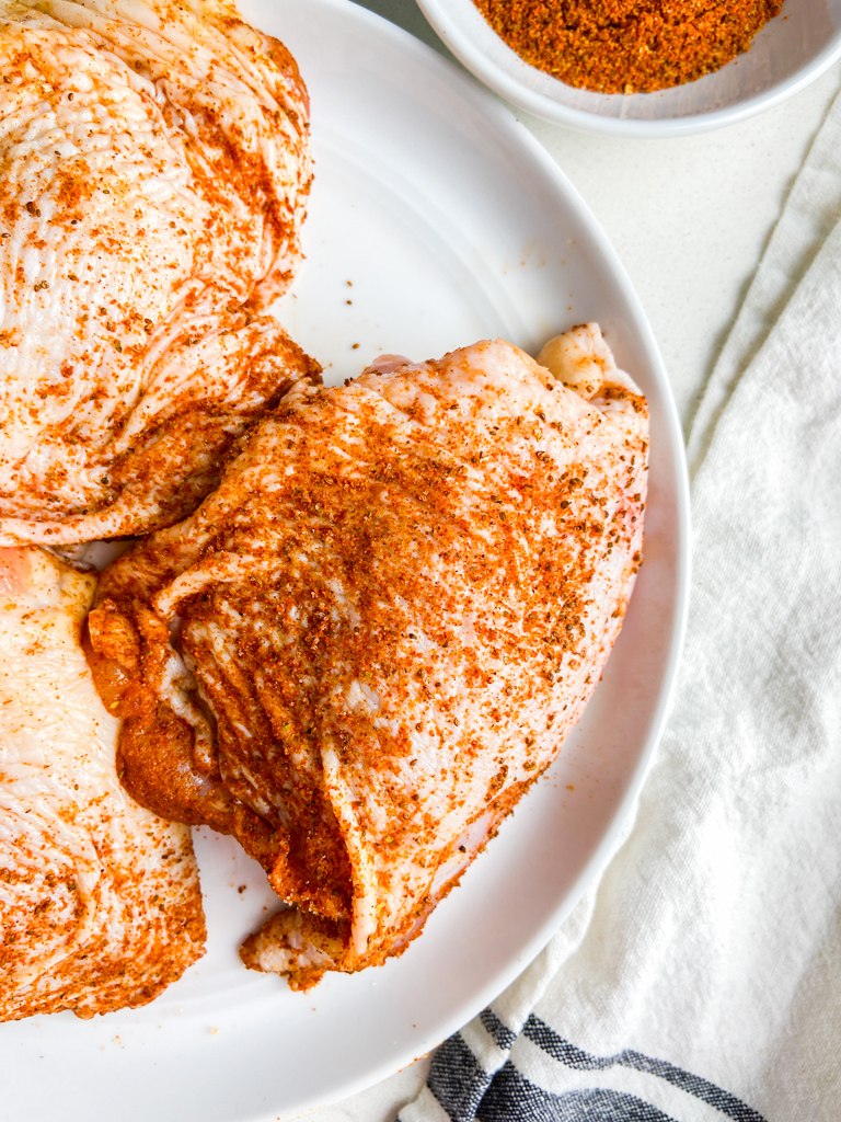 Raw chicken thigh rubbed with old bay seasoning on a white plate. 