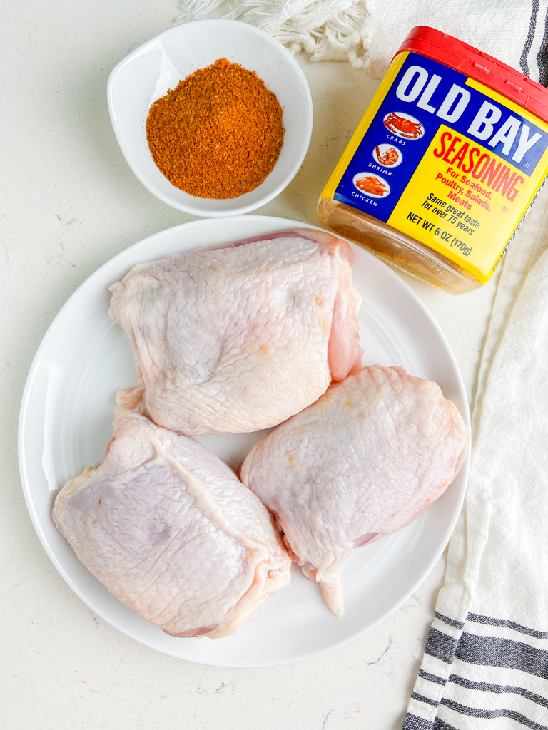 Overhead photo of raw chicken thighs on a white plate with old bay seasoning. 