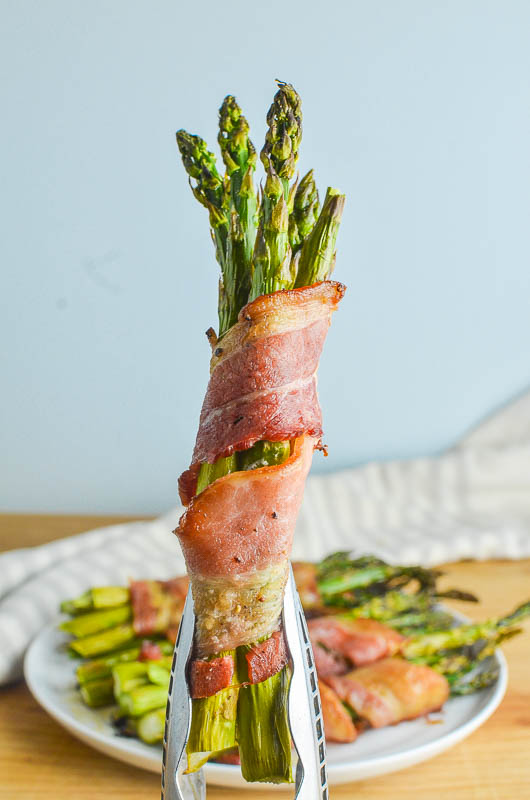 Asparagus wrapped bacon bundle held in kitchen tongs with blue background.