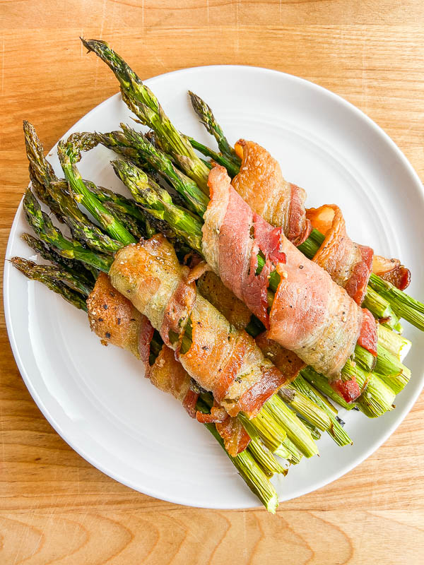 Overhead photo of bacon wrapped asparagus on a white plate and wooden cutting board. 