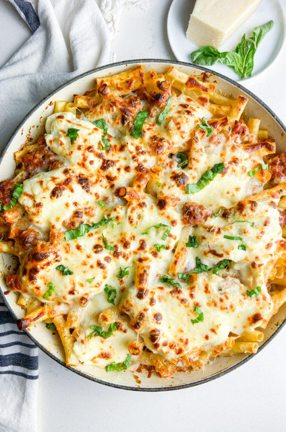 Overhead photo of baked ziti in a skillet with a stripped towel and parmesan cheese.