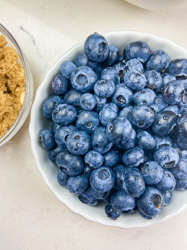Rinsed blueberries in a white bowl on a white counter. Brown sugar in a clear bowl off to the left side. 