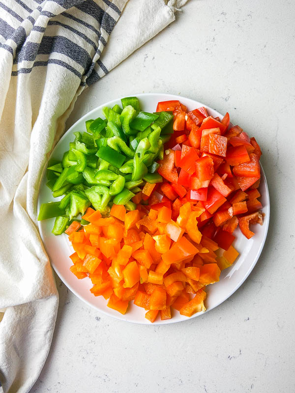 A plate of diced red, green and orange bell peppers on a white background with a striped towel. 