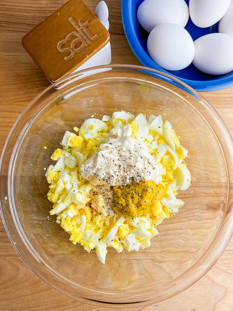 chopped eggs, mayonnaise, mustard, salt, pepper and garlic in a glass bowl on a wooden cutting board. 