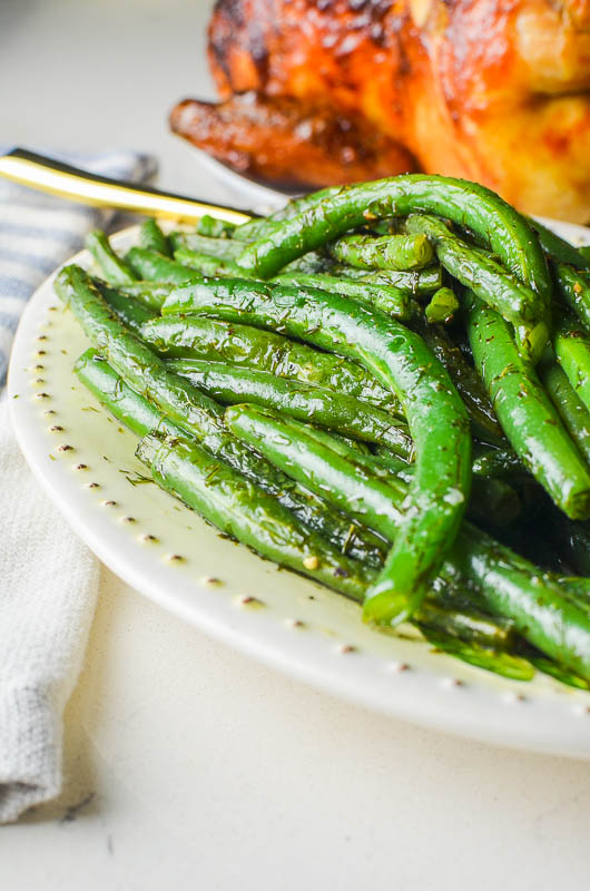 Side view photo of green beans on an oval plate with a roasted chicken in the background. 