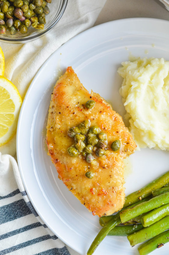 Chicken piccata on a white plate with green beans and mashed potatoes.
