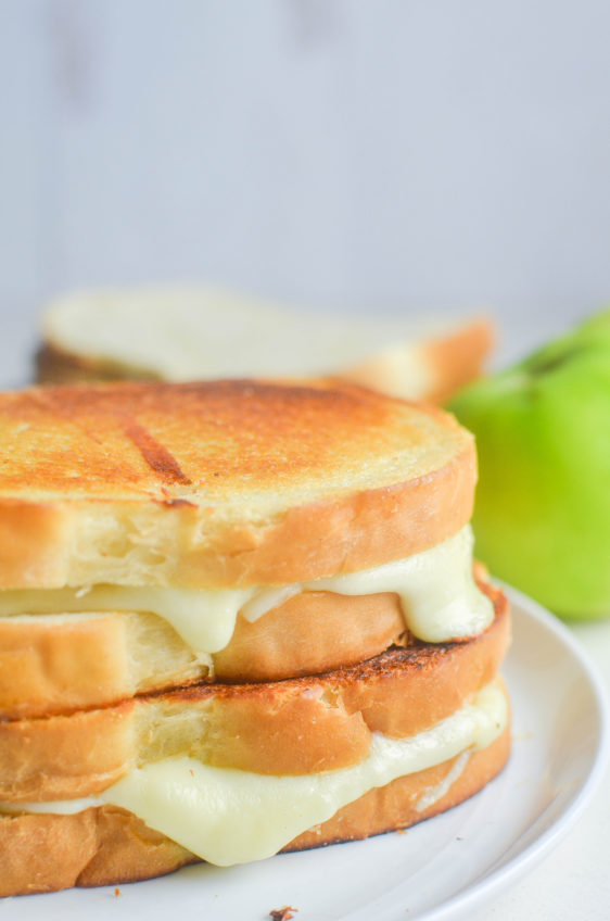 Two fried green tomato grilled cheese sandwiches stacked on each other on a white plate with a green tomato in the background.