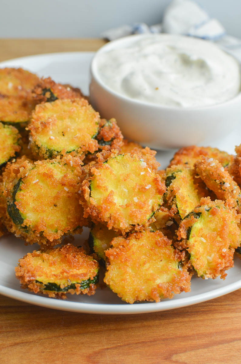 A white plate with fried zucchini and ranch dip on a wooden cutting board. 