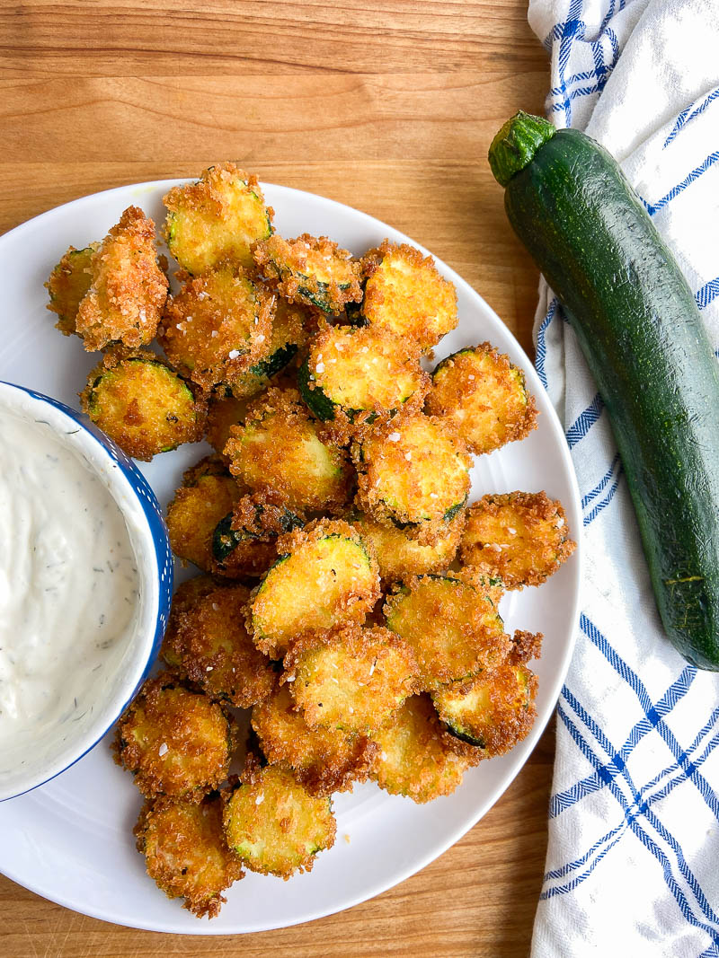 Overhead photo of fried zucchini on a white plate with uncut zucchini on a white and blue towel. 