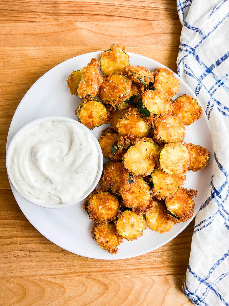 Fried Zucchini on a white plate with ranch dip.  A wooden cutting board and a blue and white napkin. 