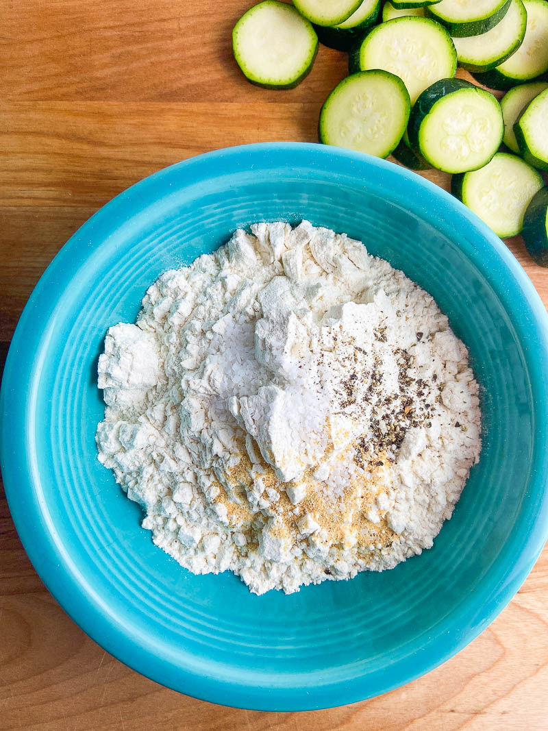 Seasoned flour in a teal bowl with zucchini on a wooden cutting board. 