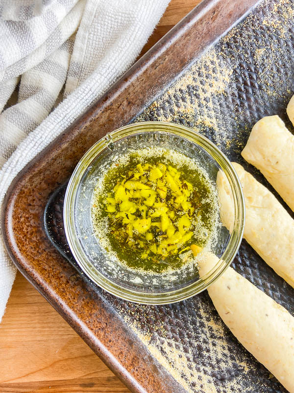 Garlic Butter in a clear bowl next to breadsticks on a baking sheet. 