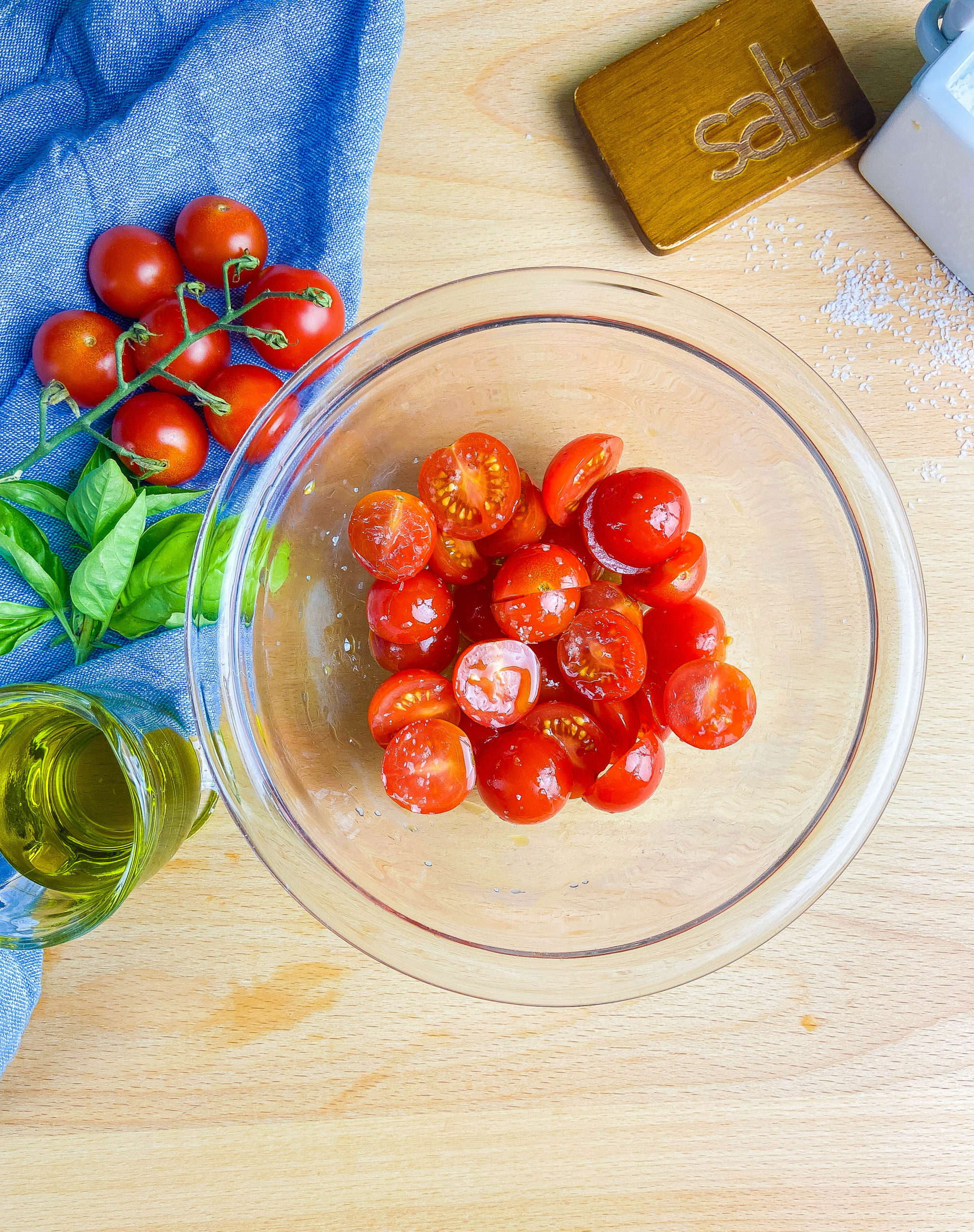 Overhead photo of cherry tomatoes in a glass bowl on a wooden cutting board. Olive oil, basil and tomatoes on a blue towel.
