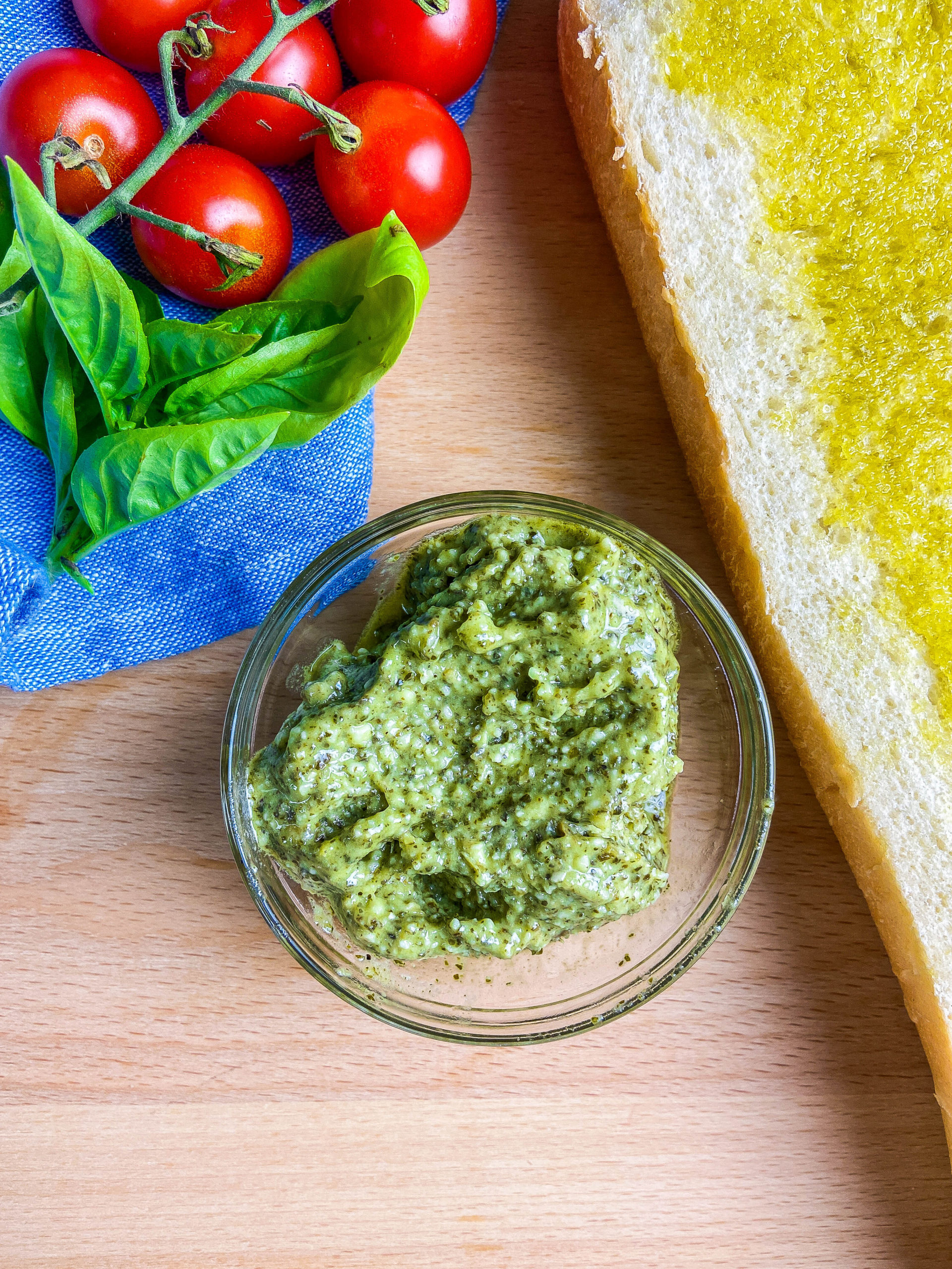 Overhead photo of pesto in a clear glass bowl. Loaf of bread, blue towel, basil and tomatoes on a wooden cutting board. 