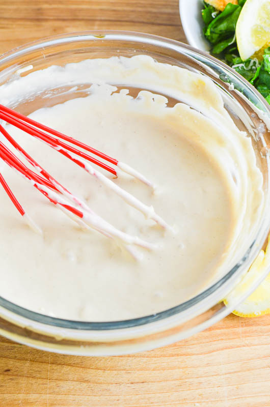 A red whisk whisking caesar dressing in a glass bowl. 