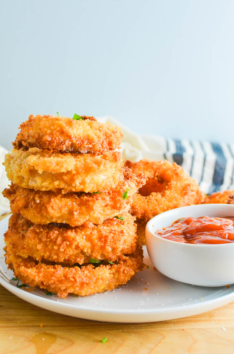 A stack of fried onion rings on a white plate with ketchup and light blue background. 