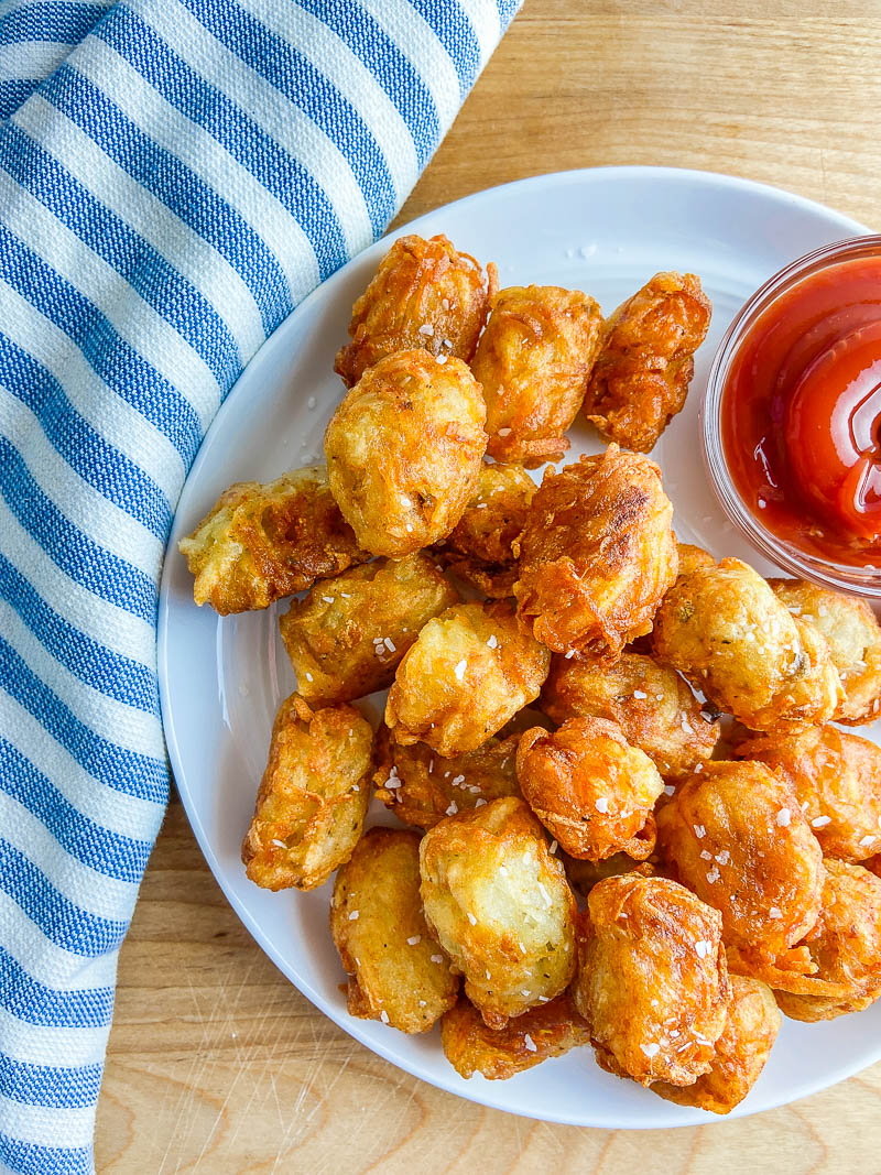 Tater tots on a white plate on a wooden cutting board with striped towel.