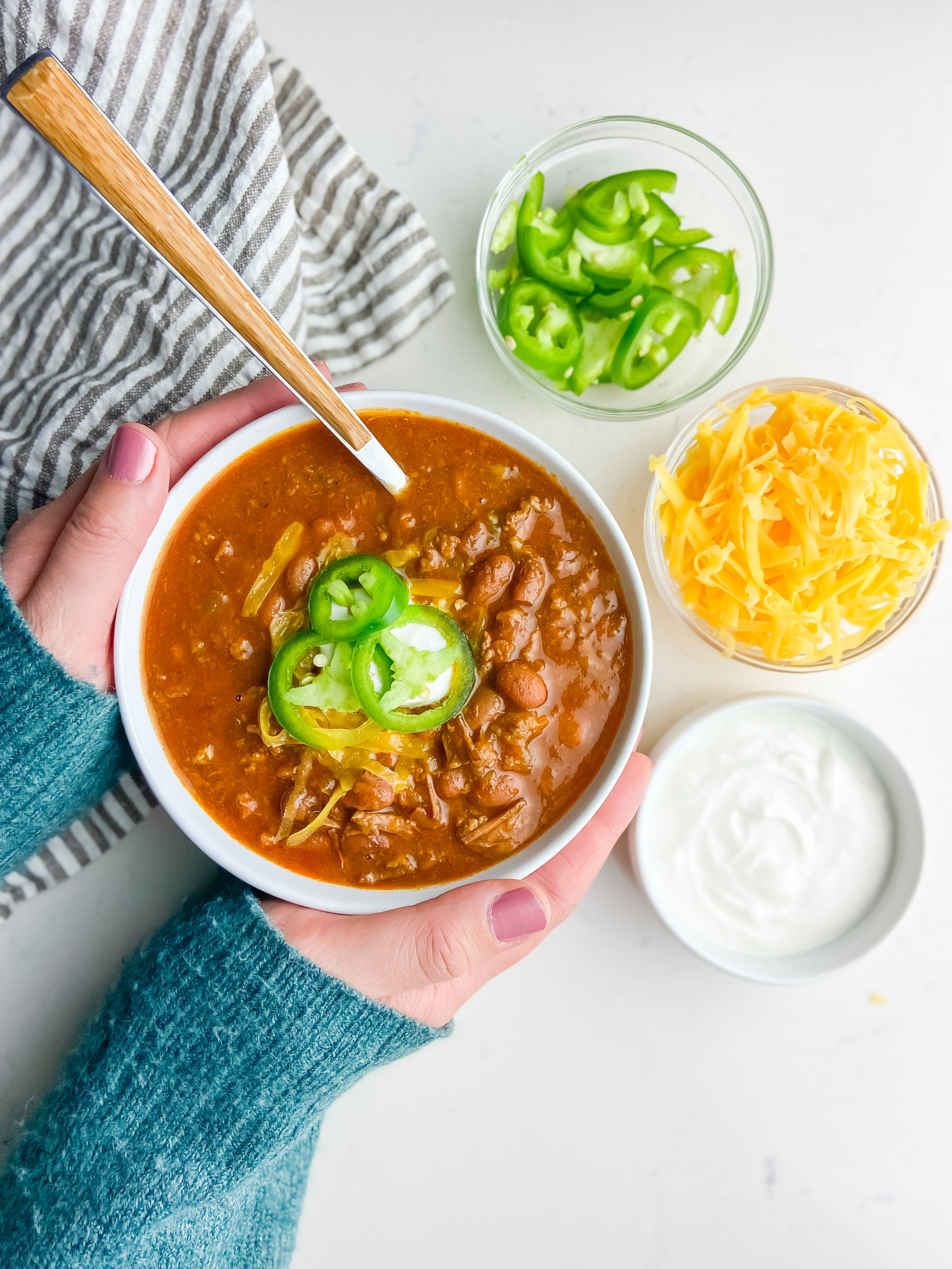 Hands holding a bowl of chili with toppings on the side. 