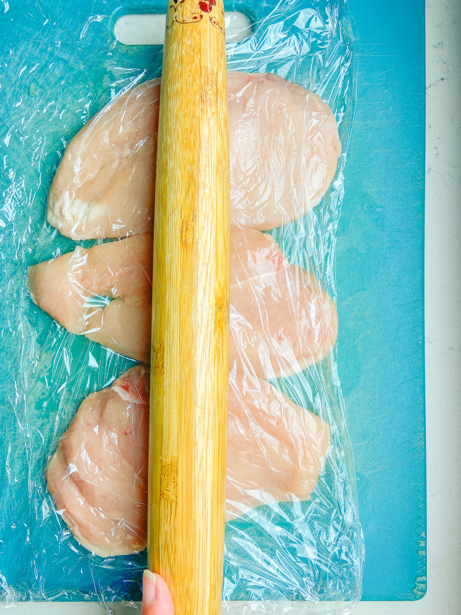 Chicken breasts between two pieces of plastic wrap on a teal cutting board with a rolling pin. 