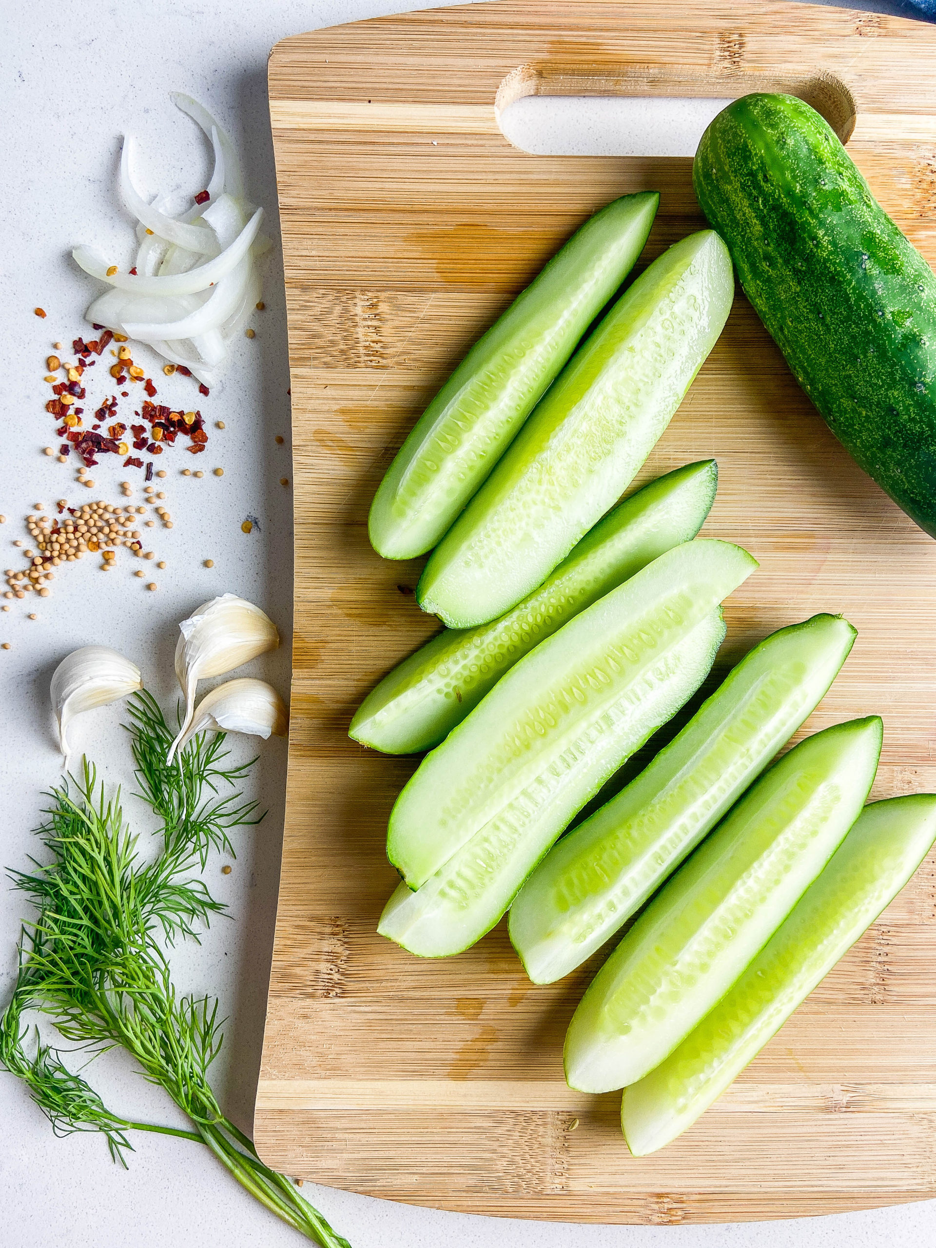 Cucumber spears on a wooden cutting board with dill, garlic cloves, mustard seeds and crushed red peppers. 