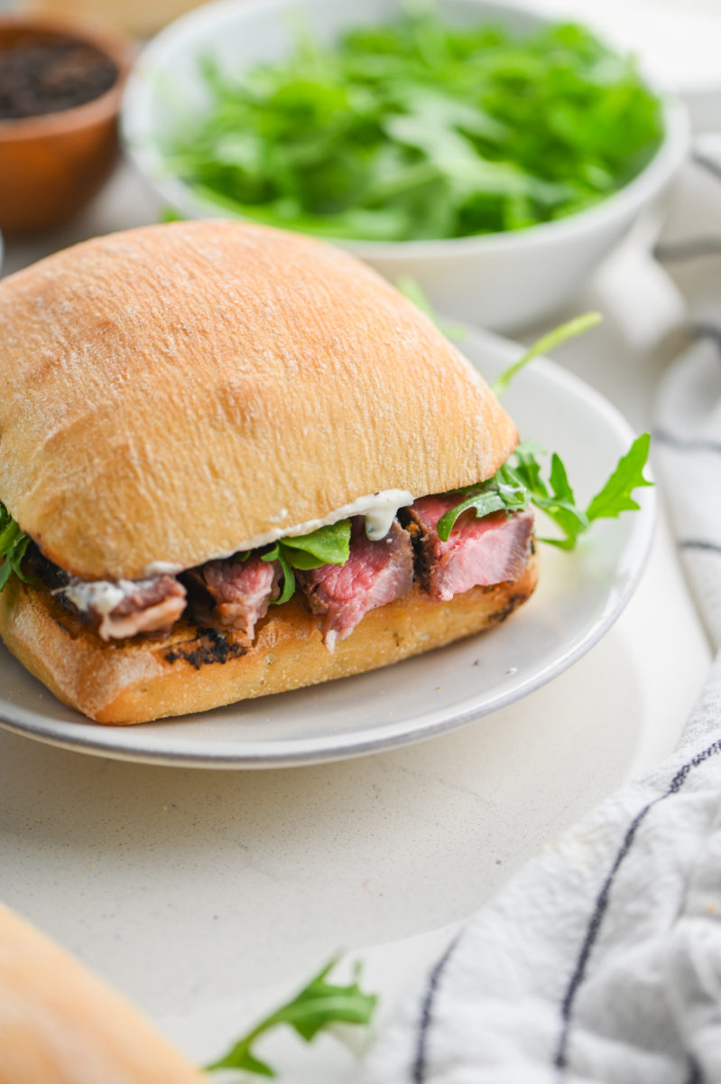 Ribeye steak sandwich on a white plate with arugula.