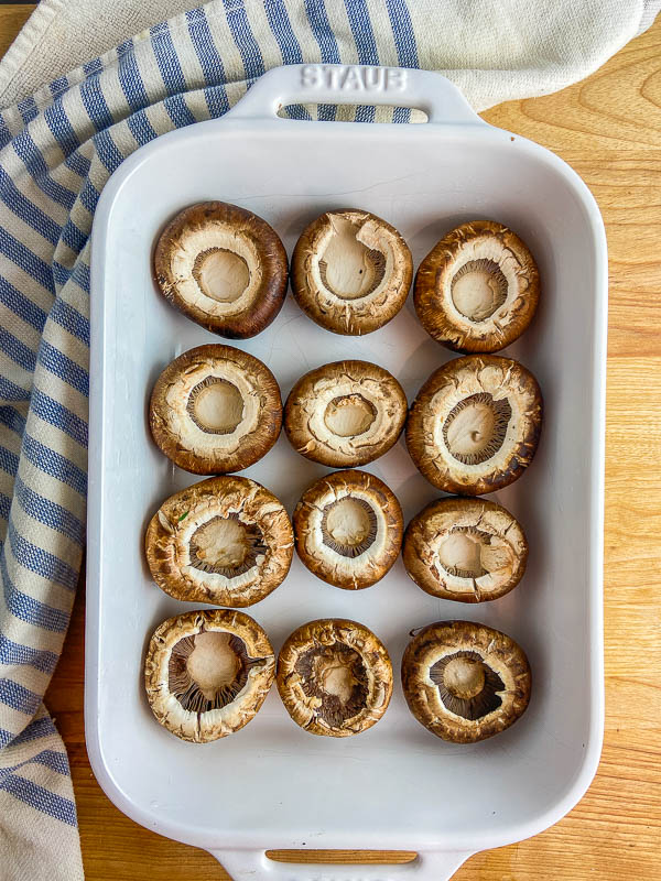 Mushroom caps in a white casserole dish with a striped towel and cutting board. 