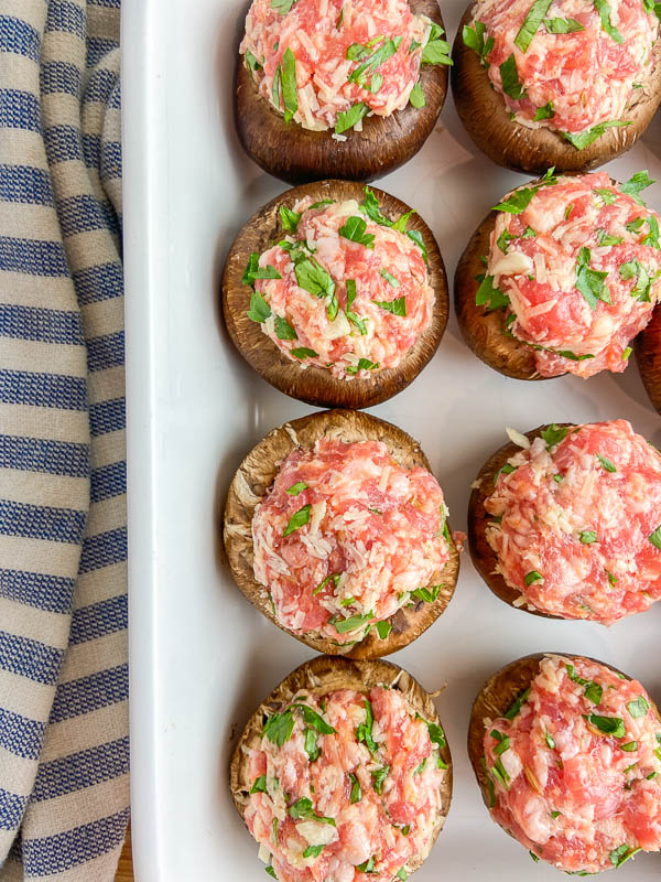 Mushrooms stuffed with raw sausage in a white casserole dish with a striped napkin. 