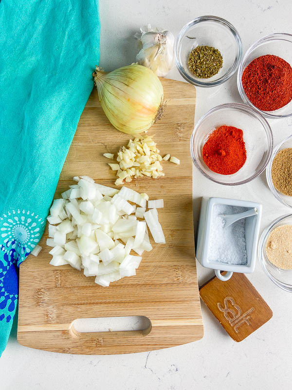 Ingredients for seasoned taco meat. Chopped onions and garlic on a cutting board with chili powder, salt, oregano, cumin and paprika in bowls. 
