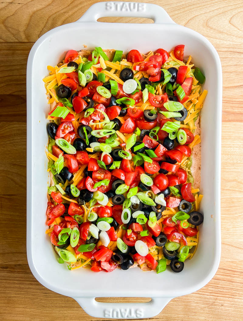 Seven layer dip in a white dish on wooden cutting board. 