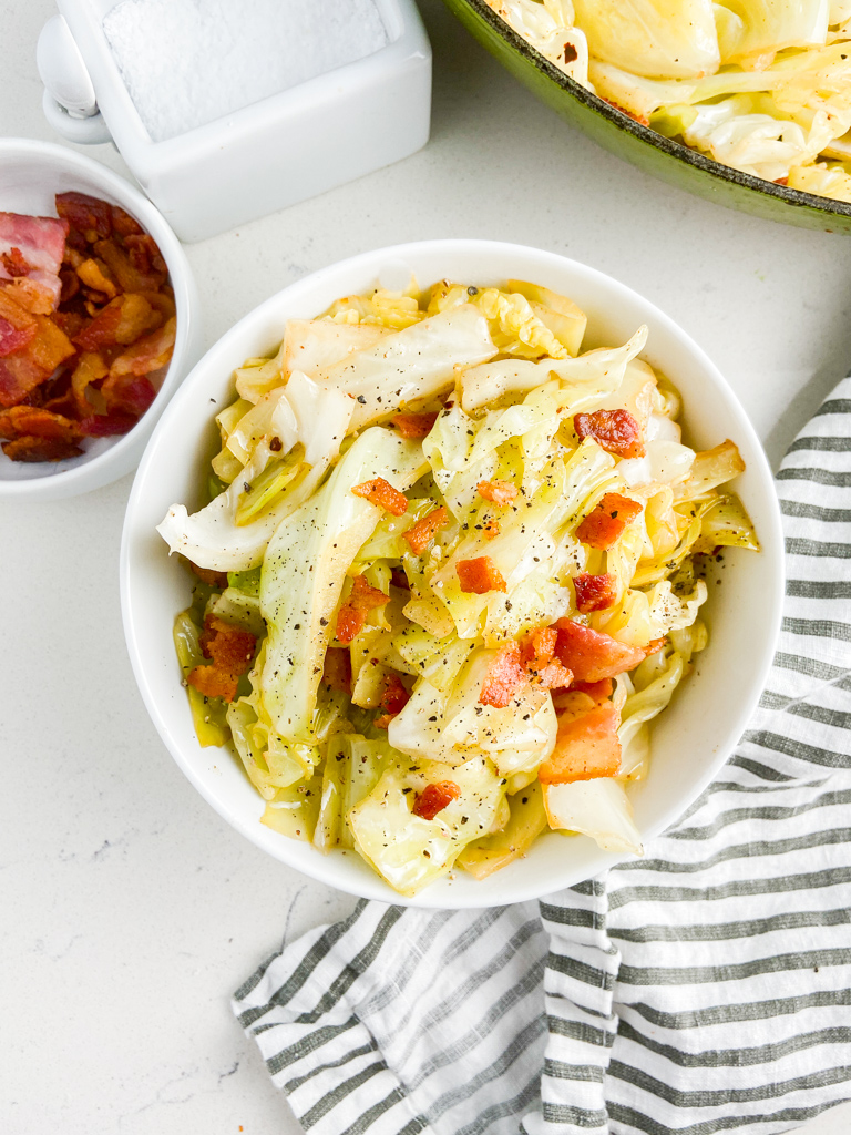 Fried cabbage in a white bowl with a striped napkin and bowl of bacon. 