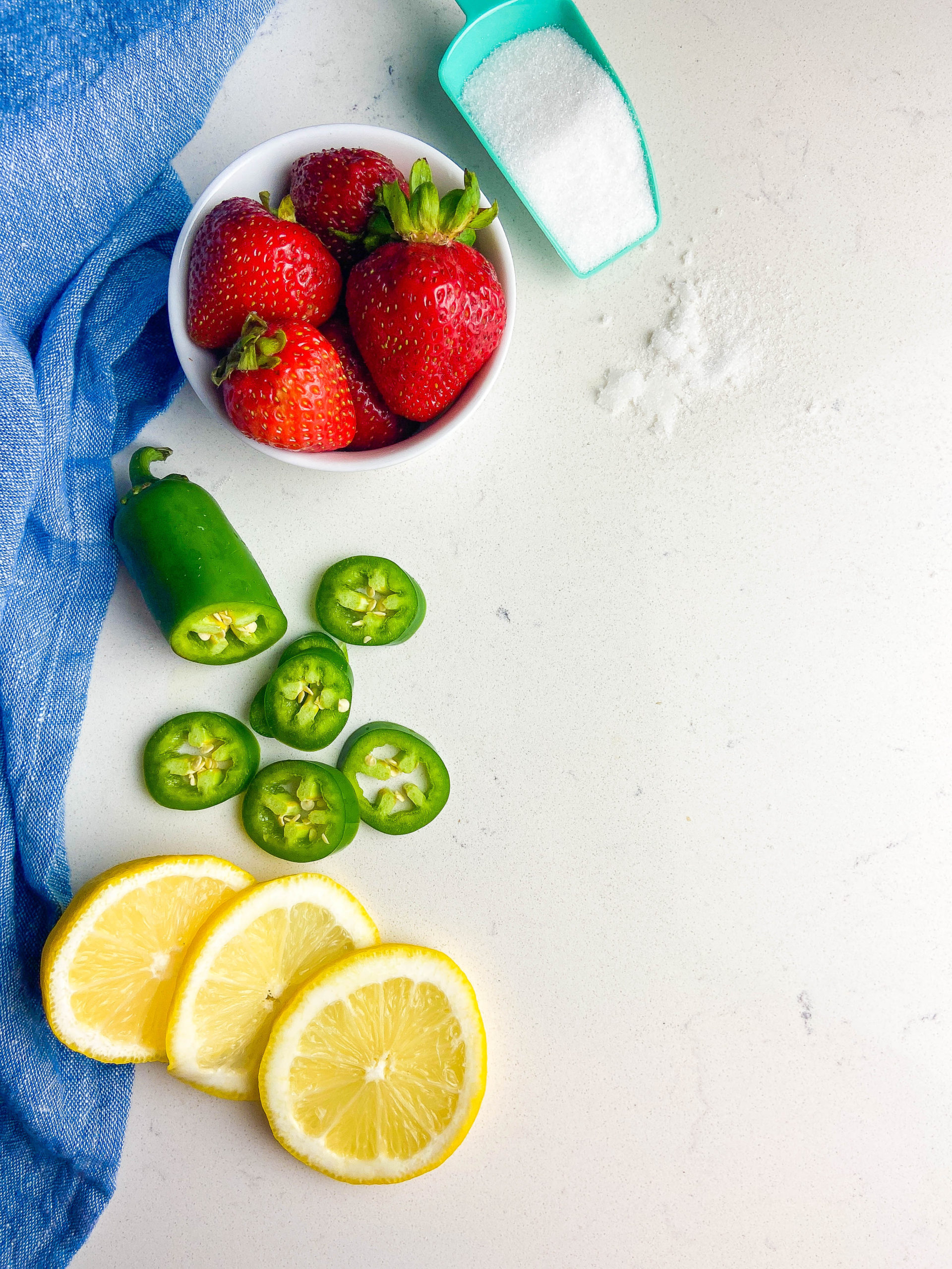 Ingredients for jalapeno jam on white surface. Slice lemons, sliced jalapenos and strawberries in a white bowl. 