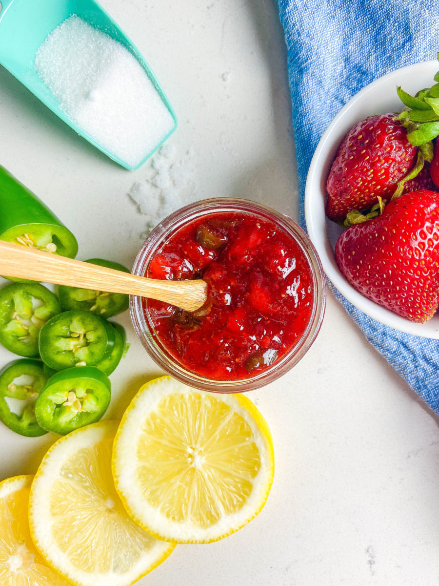 Overhead photo of strawberry jalapeno jam on white background with lemons, jalapeno, sugar and fresh strawberries