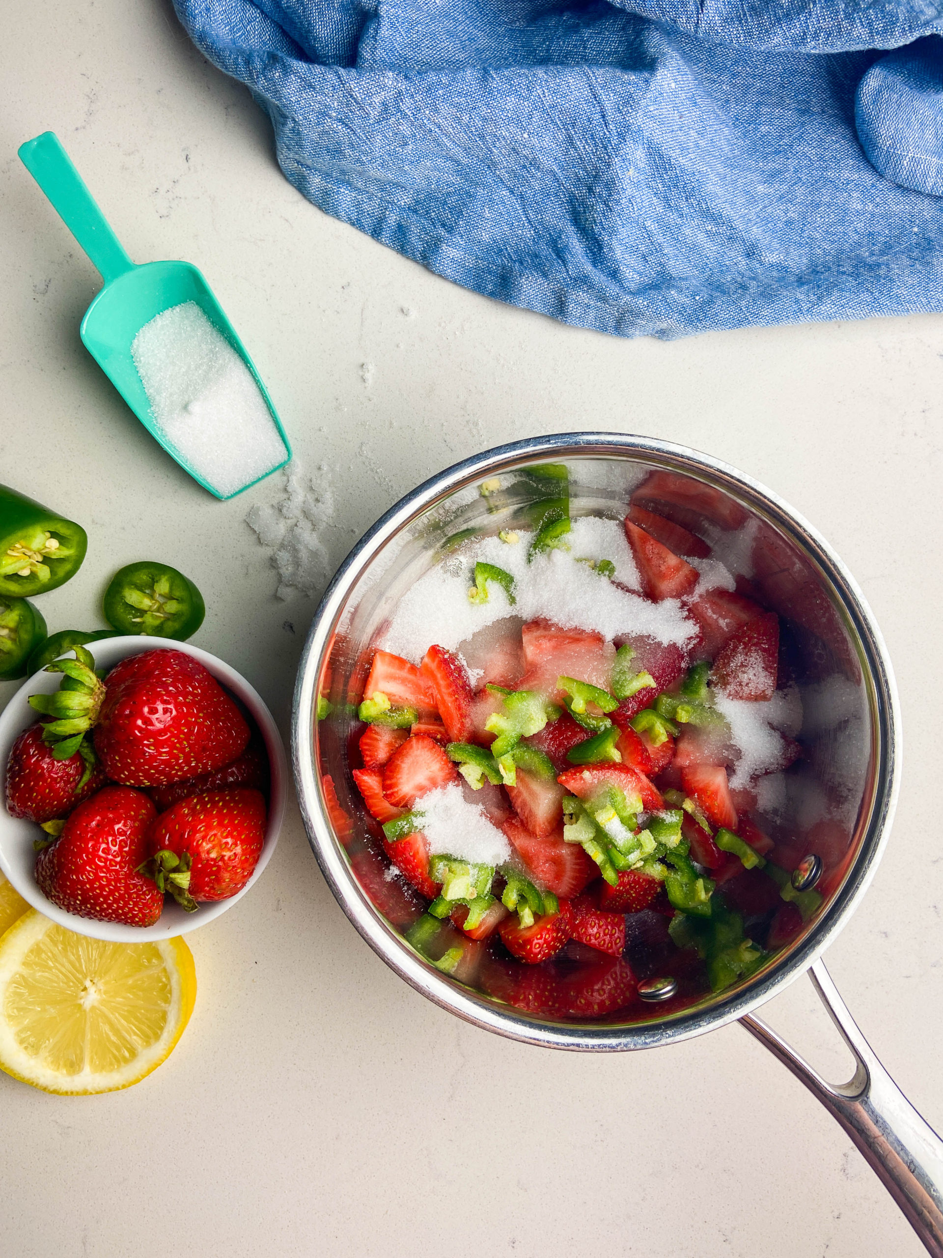 All jam ingredients in a saucepan on a white surface. Also shown: strawberries in a white bowl, scoop of sugar, lemon slices and sliced jalapenos. 