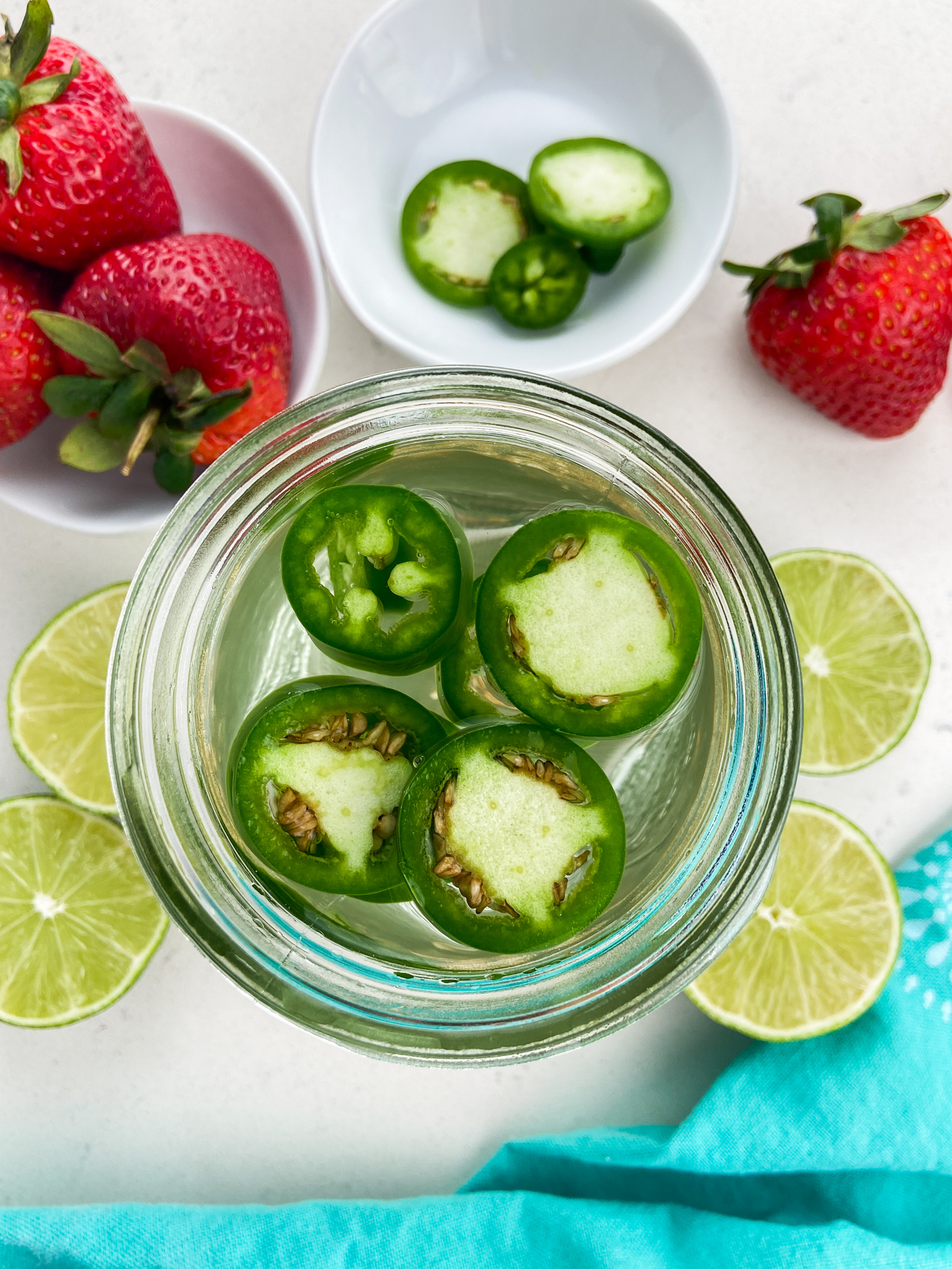 Overhead photo of jalapeños in tequila with strawberries and limes surrounding it.