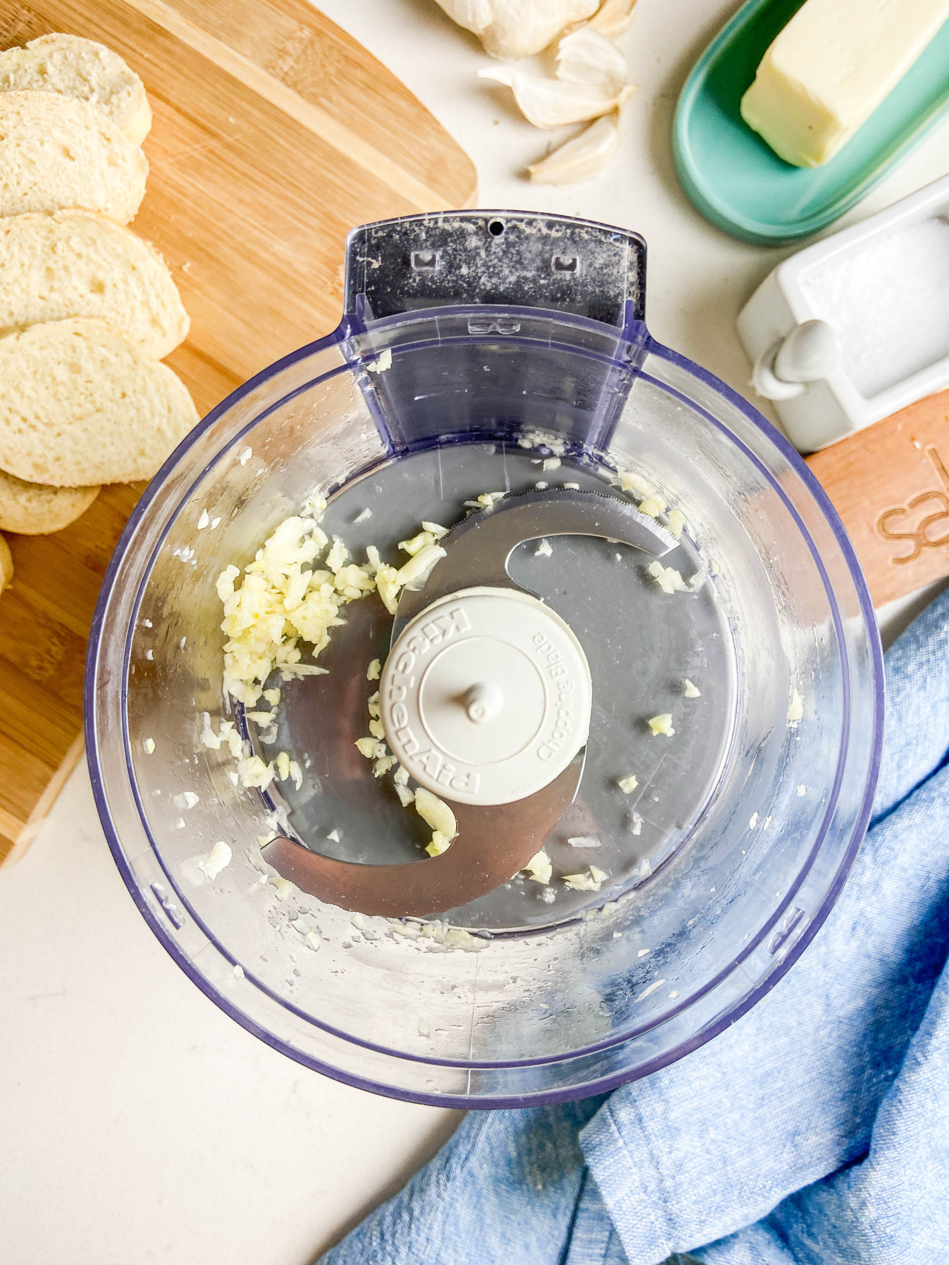 Minced garlic in a food processor. Bread, blue towel and garlic in the background. 