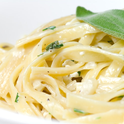 close up photo of creamy herb fettuccine in white bowl with sage leaf on top.