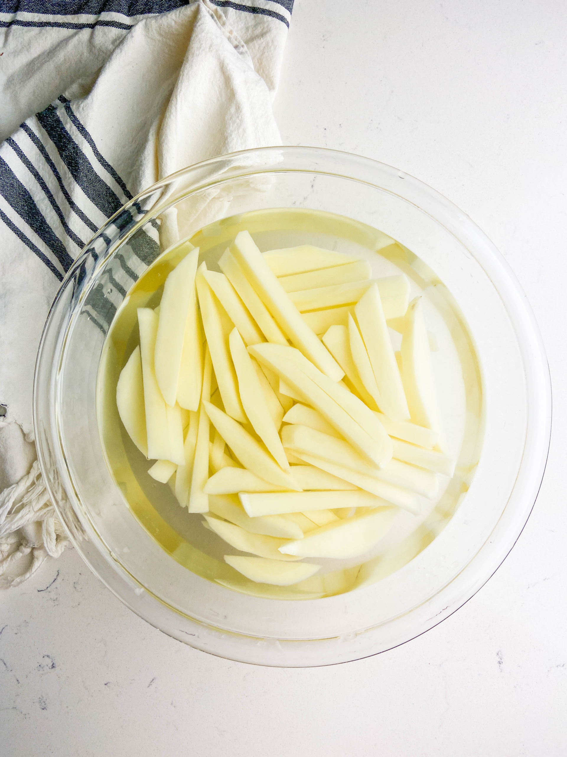 French fries soaking in cold salted water in a clear bowl on a white background. 