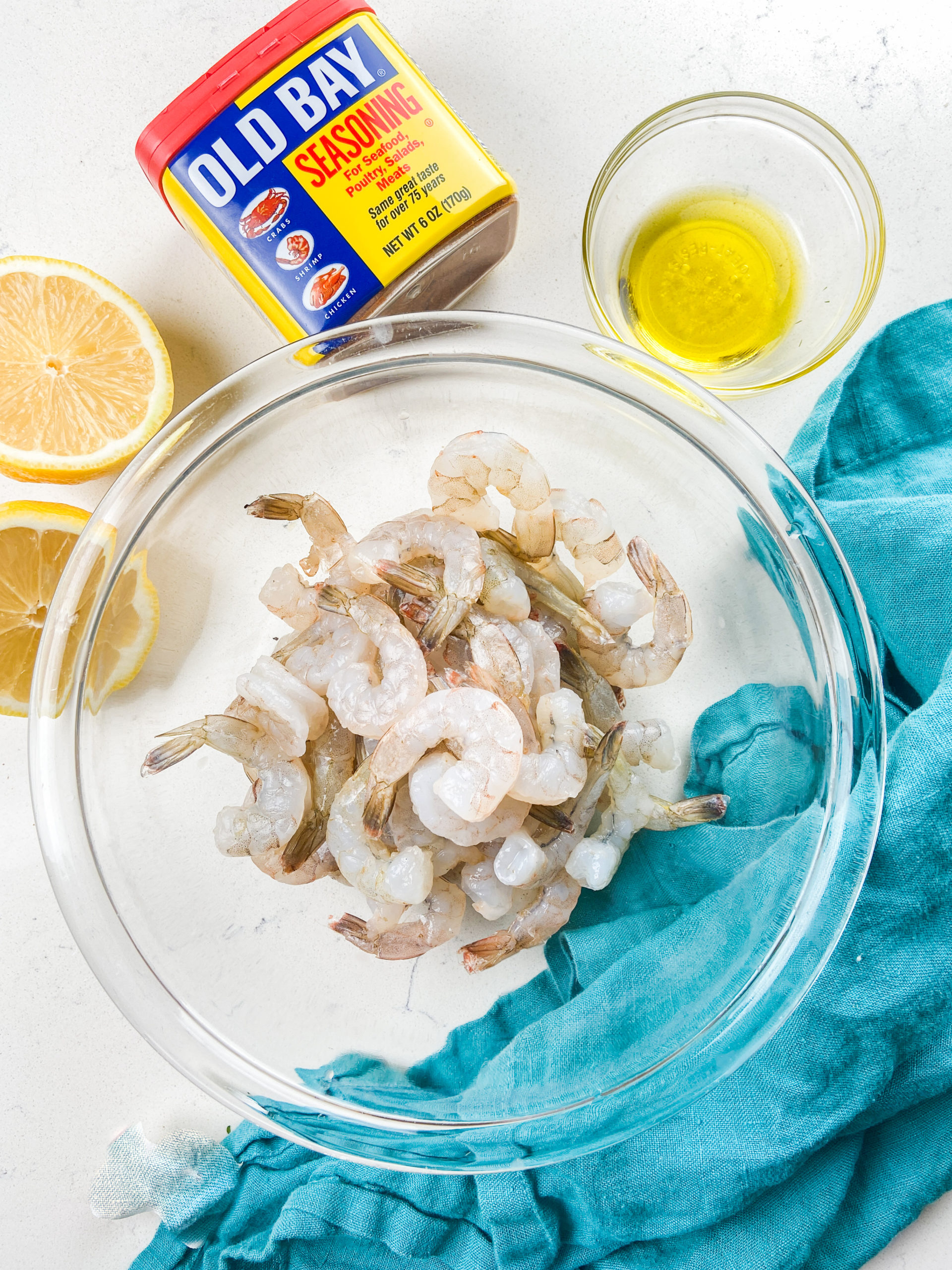 Overhead photo of raw shrimp in a glass bowl with lemon, olive oil and old bay seasoning. 