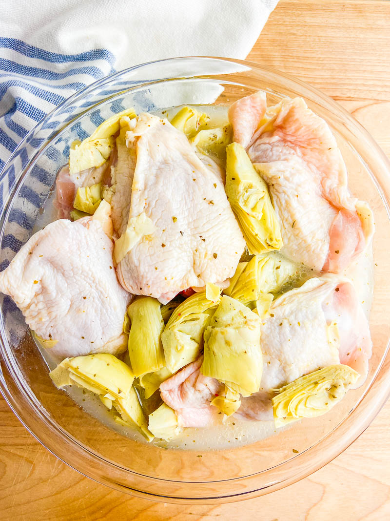 Chicken marinating with artichokes in glass bowl on cutting board with stripped napkin. 