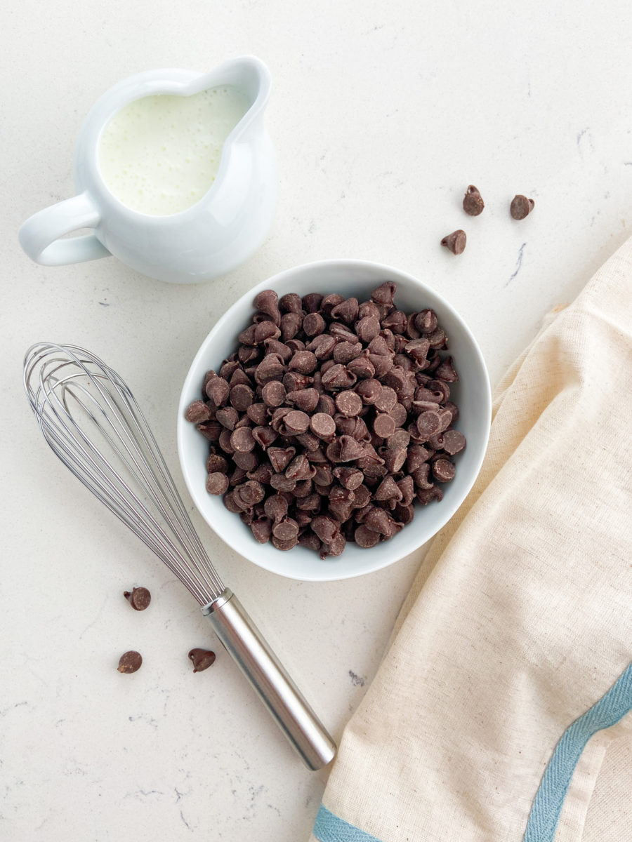 A bowl of chocolate chips, a whisk and cream. 